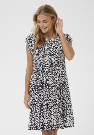 Noor naeratav naine, kes kannab lühikeste varrukatega põlvikuni ulatuvat must-valget leopardimustrilist kleiti, puudutades ühe käega juukseid.