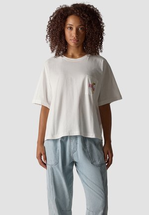 Frau mit lockigem Haar, die ein lockeres weißes T-Shirt mit einem kleinen rosa Flamingo auf der Brusttasche und lockere hellblaue Jeans trägt.