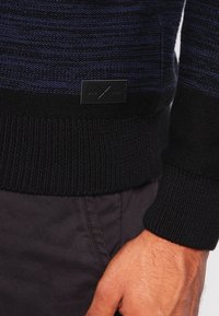 Close-up van de torso van een persoon die een zwart en marineblauw gebreid sweater draagt met een leren merkpatch boven de zoom en donkere broek.