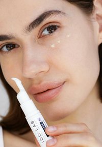 Weißes Röhrchen mit einer spitzen Applikatorspitze, beschriftet mit "IDO LAB", wird nahe dem Gesicht einer Person gehalten, die kleine Punkte Creme unter einem Auge zeigt.