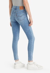 Person som bär ljusblå slim-fit jeans och vita sneakers, stående med ena handen avslappnat vid sidan, vänd bort.