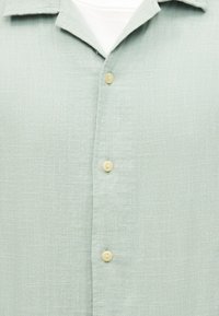 Ανοιχτοπράσινο υφασμάτινο πουκάμισο με τρεις ορατά μπεζ κουμπιά και λευκό ύφασμα κάτω από το γιακά.