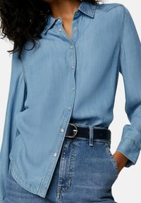 Lichtblauwe spijkerblouse met een kraag, knoopsluiting en lange mouwen. Draagt een paar donkerblauwe jeans en een zwarte riem met een zilveren gesp.