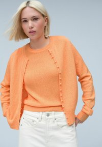 Cardigan arancione chiaro con tessuto testurizzato e dettagli pom-pom lungo la parte anteriore, indossato sopra un maglione arancione abbinato e jeans bianchi.