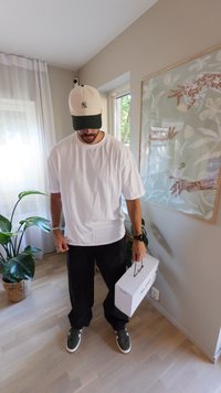 Weißes übergroßes T-Shirt, schwarze weite Hose, grüne Sneakers und eine grün-weiße Mütze. Hält eine weiße Schuhbox-Tasche. Innenraum mit Pflanzen.