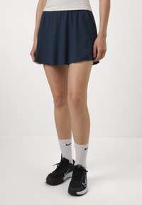 Falda atlética navy fabricada con un material liviano, que presenta una cinturilla elástica y un ligero vuelo. Combinada con zapatillas negras y calcetines blancos.