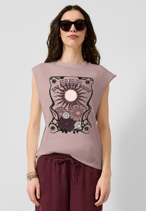 Frau mit langem, welligem Haar, die schwarze Sonnenbrillen trägt, ein ärmelloses mauvefarbenes Oberteil mit Sonnen- und Blumenmotive und bordeauxrote Hose mit Kordelzug.