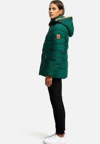 Green puffer jacket with a fur-lined hood, textured fabric, side pockets, and a brown accent on the sleeve. Black pants and white sneakers.