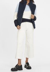 Mörkblå quiltad väst över en randig, lätt tröja i blått, krämvitt och mörkblått; kombinerat med vida vita byxor och svarta skor med grov sula.