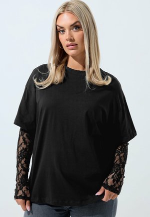 Vrouw met steil blond haar draagt een oversized zwart T-shirt en zwarte bloemen kanten lange mouwen, poserend tegen een effen lichte achtergrond.