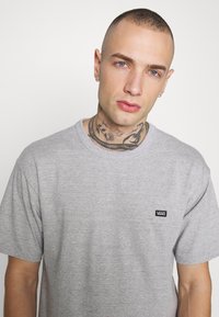 Camiseta de algodón gris con mangas cortas, cuello redondo y un pequeño logo negro de Vans en el pecho. Textura suave, corte relajado.