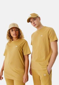 Deux modèles portent des t-shirts en coton beige à manches courtes avec un petit logo. Une casquette beige avec un logo complète les tenues.