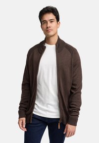 Brązowy kardigan na zamek błyskawiczny z prążkowanymi detalami na ramionach, teksturowana tkanina i beżowy uchwyt zamka, noszony na białej koszuli i niebieskich dżinsach.
