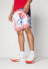 Pantalones cortos blancos con un gran logotipo de los Yankees en rojo y azul, que presenta patrones de costura de béisbol. Llevados con zapatillas Nike blancas y naranjas.