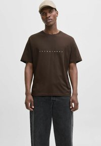 Brun bomulds t-shirt med korte ærmer og rund hals, med "JACK&JONES" trykt i hvid på brystet. Båret med sorte jeans.