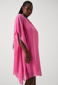 Pink, flowy dress made of lightweight fabric with a V-neck and wide sleeves, featuring a hem that curves upwards on the sides.