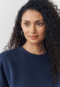 Navy plava majica s visokim okruglim izrezom, izrađena od mekanog materijala. Model ima kovrčavu kosu i nosi male naušnice u obliku obruča.