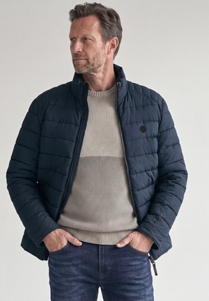 Mann trägt eine marineblaue gesteppte Jacke über einem beigen Pullover und dunklen Jeans, steht mit den Händen in den Taschen und schaut nach links.
