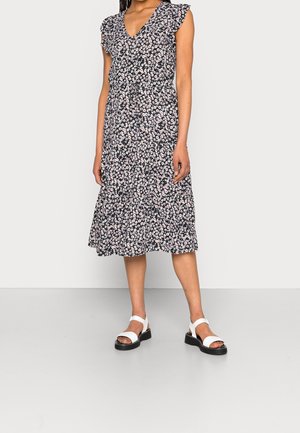 Kvinde iført en sort midikjole med hvidt og pink blomsterprint og hvide sandaler med åben tå, stående på en ensartet lys baggrund.
