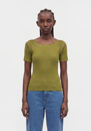 Femme aux cheveux tressés portant un haut côtelé ajusté vert olive à manches courtes et un jean bleu taille haute, debout devant un fond uni.