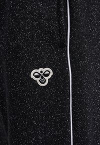 Schwarzer Strickstoff mit silbernen Flecken, ausgestattet mit einem weißen Bienenlogo und kontrastierenden weißen Paspelierungen an der Seite. Weiche Textur, lässiges Design.