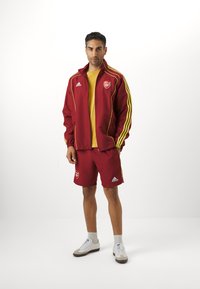Rød sportsjakke med gule detaljer og logoer, matchende røde shorts og hvite sneakers. Modellen står oppreist med et nøytralt uttrykk.