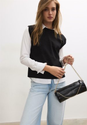 Młoda kobieta w białej koszuli i czarnym bezrękawniku, trzymająca czarną cylindryczną torebkę na złotym łańcuszku.