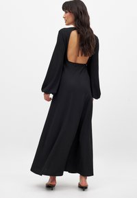 Abito maxi nero con maniche lunghe a palloncino e schiena scoperta. Realizzato in tessuto testurizzato, presenta un design fluido che si estende fino al pavimento.