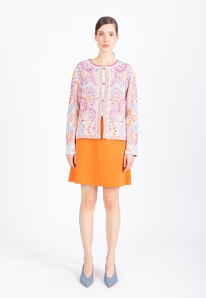 Naine seisab, kandes mitmevärvilise lillemustriga kardigani, erkoranži seelikut ja helesiniseid terava ninaga kingi valgel taustal.