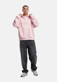 Sweatshirt à capuche rose avec une poche avant, assorti à un jean ample gris foncé et des baskets grises, sur fond uni.