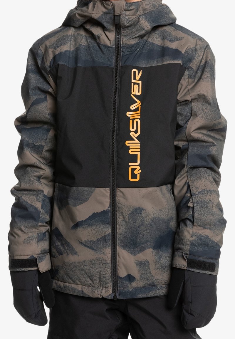 Enfant portant une veste d'hiver à motif camouflage avec un panneau noir sur la poitrine et un logo jaune vertical "Quiksilver", des gants noirs et un pantalon noir.