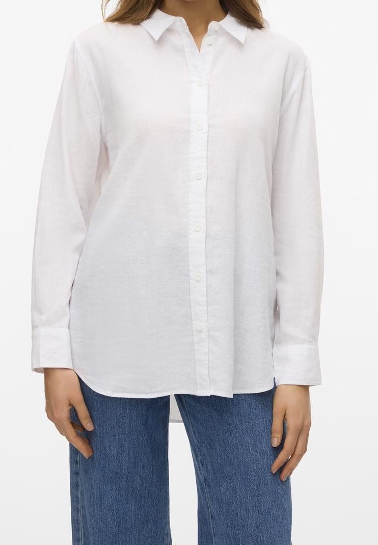 Femme portant une chemise blanche à manches longues boutonnée et un jean en denim bleu, debout les bras détendus le long du corps.