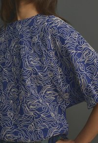Modrý vršek z látky s bílým vlnitým vzorem, výstřihem ve tvaru člunu a volným střihem s krátkými rukávy. Materiál se zdá být lehký a prodyšný.
