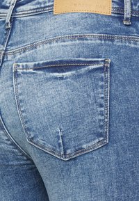 Jeans de ganga azul skinny com um bolso traseiro com borda costurada e um patch de marca em couro acima. Textura suave com áreas desbotadas.