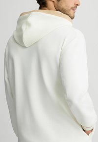 Hoodie in Off-White mit beigem Innenfutter. Verfügbar mit weicher Textur und lockerer Passform, großem Kapuzenkragen und Kängurutasche.