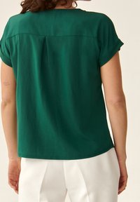 Vihreä paita käärittyine hihoineen, pehmeä kangas ja takana laskostus. Alahelma on kaareva, käytetään vaaleiden housujen kanssa.