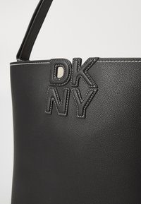 Melna ādas somiņa ar izvirzītu "DKNY" logo, balto šuvju un strukturētu formu. Gluda tekstūra ar minimālistisku dizainu.