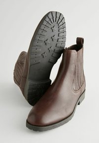 Botine din piele maro, cu un panou elastic nervurat și tab pentru a fi tras de pe picior, având o talpă din cauciuc texturat negru și vârful rotunjit.