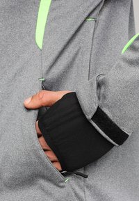 Veste en polaire grise avec une poche avant, des poignets noirs et des accents verts. Le tissu a une texture lisse et un design sportif.