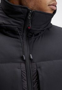 Doudoune noire avec un col montant, équipée d'une fermeture éclair frontale avec un tirette en forme de feuille rouge, et des cordons élastiques au niveau du cou.