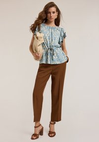 Chemise bleue à motifs avec des manches volantes, pantalon marron taille haute et sandales à talons marron. Le mannequin tient un sac beige.