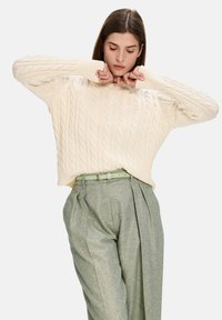 Chunky cream cable-knit sweater with a relaxed fit and crew neckline, paired with textured grey plaid trousers and a light green belt.