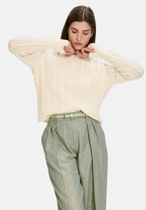 Flauschiger, cremefarbener Strickpullover mit entspannter Passform und Rundhalsausschnitt, kombiniert mit strukturierten grauen Karottenhosen und einem hellgrünen Gürtel.