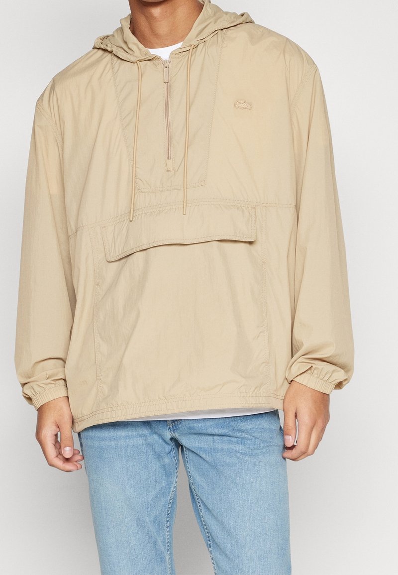Homme portant une veste à capuche beige avec demi-fermeture éclair et poche avant, associée à un jean bleu clair.