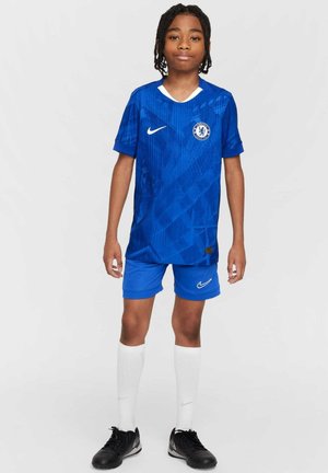 Junge Person steht, trägt ein blaues Chelsea-Fußballtrikot mit weißen Socken und schwarzen Fußballschuhen, vor einfachem hellem Hintergrund.