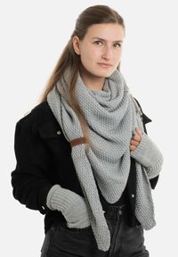Grijze gebreide sjaal elegant gedrapeerd, met een gestructureerde afwerking. Gecombineerd met grijze gebreide vingerloze handschoenen, tegen een zwarte ribfluwelen jas.