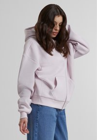 Felpa con zip di colore rosa chiaro, dal taglio comodo, con ampie tasche e polsini a coste. Il tessuto appare morbido e ha una consistenza liscia.