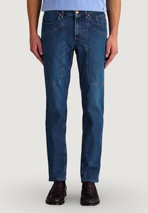 Hombre vistiendo jeans azul oscuro de corte recto con grandes bolsillos delanteros y mocasines marrón oscuro, de pie frente a un fondo sencillo.