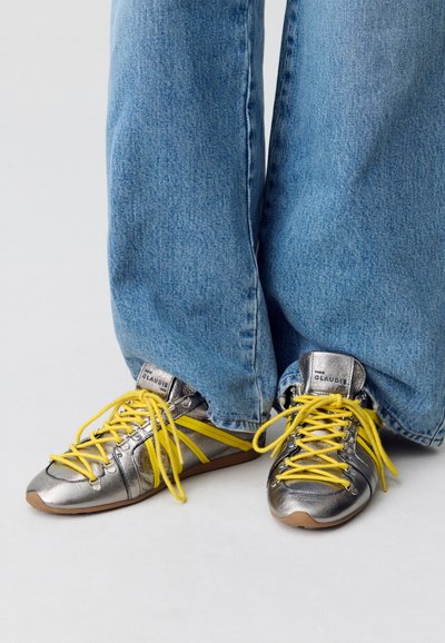 Sneakers metallici argento con lacci e dettagli gialli, caratterizzati da una punta arrotondata e una suola imbottita, abbinati a jeans di denim azzurro chiaro.