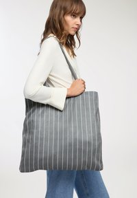 Szara torba na ramię z białymi pionowymi paskami, wykonana z tkaniny. Duży, prostokątny kształt z mocnymi, długimi uchwytami do łatwego noszenia.